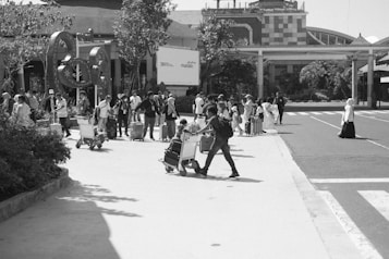 A busy outdoor scene at an airport or transit hub with a large group of people, many with luggage and carts, moving across a paved area. Buildings and landscaping are visible in the background, with people appearing to be in transit or awaiting transport.