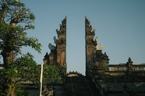 An ornate split gate, constructed from stone, features intricate carvings and is flanked by lush greenery. The clear blue sky forms the backdrop, enhancing the traditional architectural aesthetics.