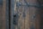 An aged wooden door with heavy, ornate metalwork and intricate patterns. The metal handle and lock show signs of wear, adding to the vintage appearance. The door is composed of vertical wooden planks with iron bolts securing them.
