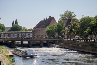 A scenic river scene featuring a modern riverboat with a glass roof navigating through a waterway. The river is flanked by charming historic buildings with steep roofs and surrounded by lush green trees. A small bridge with potted plants and pedestrians spans the river.