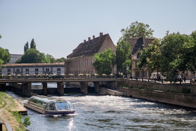 A scenic river scene featuring a modern riverboat with a glass roof navigating through a waterway. The river is flanked by charming historic buildings with steep roofs and surrounded by lush green trees. A small bridge with potted plants and pedestrians spans the river.