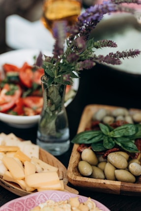 A close-up of a variety of appetizer dishes including a wooden platter with olives, sun-dried tomatoes, and basil leaves. Nearby is a board with assorted cheese slices and a bowl of fresh salad featuring tomatoes and greens. A small vase with purple flowers adds a decorative touch.