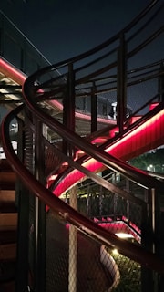 Curving architectural structure featuring metal railings and mesh panels, illuminated with red and white lights against a dark night sky.