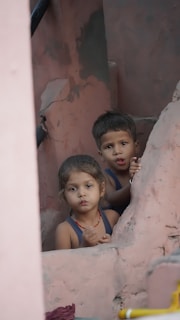 Two young children appear to be playing or exploring among pink, weathered structures, looking directly at the viewer with curious expressions.