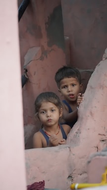 Two young children appear to be playing or exploring among pink, weathered structures, looking directly at the viewer with curious expressions.