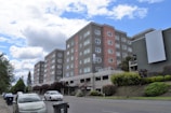 A multi-story residential building with a modern design is surrounded by greenery, with cars parked alongside the road in an urban setting. The building features a combination of gray and pinkish tones and is set against a partly cloudy sky.