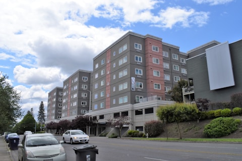 A multi-story residential building with a modern design is surrounded by greenery, with cars parked alongside the road in an urban setting. The building features a combination of gray and pinkish tones and is set against a partly cloudy sky.