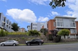 Modern townhouses with large windows and varied facades are positioned on a gentle hillside. In the foreground, two parked cars are on the street lined with small shrubs and trees. The sky is blue with scattered white clouds.