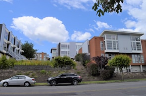 Modern townhouses with large windows and varied facades are positioned on a gentle hillside. In the foreground, two parked cars are on the street lined with small shrubs and trees. The sky is blue with scattered white clouds.