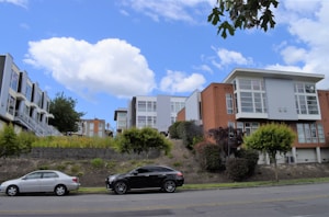 Modern townhouses with large windows and varied facades are positioned on a gentle hillside. In the foreground, two parked cars are on the street lined with small shrubs and trees. The sky is blue with scattered white clouds.