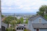 A residential area featuring houses with sloped roofs, surrounded by greenery and trees. In the background, there is a view of a distant cityscape with hills and a cloudy sky. A small parking lot with several cars and lined trash bins is visible, bordered by fences and power lines.