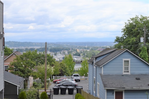 A residential area featuring houses with sloped roofs, surrounded by greenery and trees. In the background, there is a view of a distant cityscape with hills and a cloudy sky. A small parking lot with several cars and lined trash bins is visible, bordered by fences and power lines.
