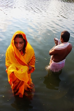 A woman in a vibrant yellow sari stands in shallow water, looking contemplative. Beside her, a man dressed in traditional attire performs a ritual with hands in prayer position, both surrounded by ripples in the water.