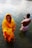 A woman in a vibrant yellow sari stands in shallow water, looking contemplative. Beside her, a man dressed in traditional attire performs a ritual with hands in prayer position, both surrounded by ripples in the water.
