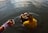 A woman partially submerged in water is performing a ritual. She is wearing a yellow and orange saree with intricate patterns, and holding a basket filled with coconuts and other offerings. Nearby, another hand adorned with colorful bangles holds a small cup, also over the water.