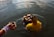 A woman partially submerged in water is performing a ritual. She is wearing a yellow and orange saree with intricate patterns, and holding a basket filled with coconuts and other offerings. Nearby, another hand adorned with colorful bangles holds a small cup, also over the water.