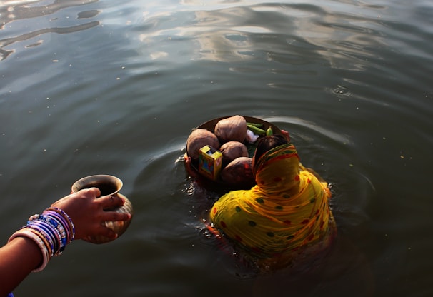 A woman partially submerged in water is performing a ritual. She is wearing a yellow and orange saree with intricate patterns, and holding a basket filled with coconuts and other offerings. Nearby, another hand adorned with colorful bangles holds a small cup, also over the water.