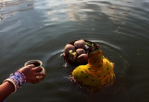 A woman partially submerged in water is performing a ritual. She is wearing a yellow and orange saree with intricate patterns, and holding a basket filled with coconuts and other offerings. Nearby, another hand adorned with colorful bangles holds a small cup, also over the water.