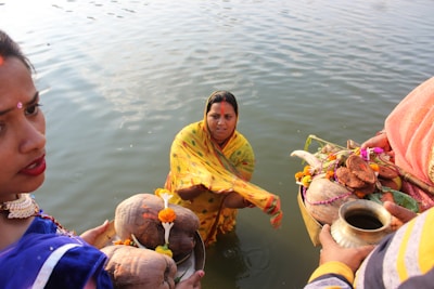 A woman stands partially submerged in water, wrapped in a yellow sari, partaking in a traditional ceremony. Another individual wearing a blue sari appears beside her, holding a ritual plate adorned with coconuts, flowers, and other ceremonial items. The water reflects the light, adding to the serene atmosphere.