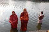 Three people stand in water near a riverbank. Two women are wearing red sarees with traditional designs and are partially submerged in the water. A man, draped in a white cloth, stands slightly further out and holds some items in his hands. The background shows a reflective surface of the water with sunlight creating ripples.