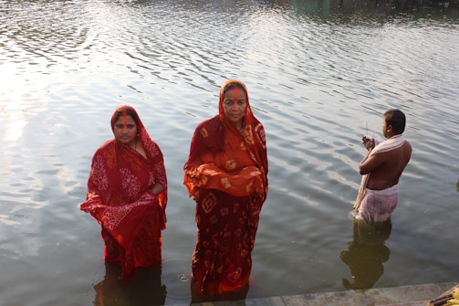 Three people stand in water near a riverbank. Two women are wearing red sarees with traditional designs and are partially submerged in the water. A man, draped in a white cloth, stands slightly further out and holds some items in his hands. The background shows a reflective surface of the water with sunlight creating ripples.