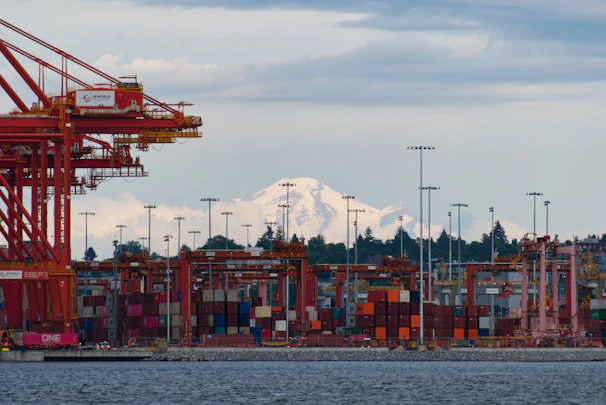 Stacked red and green maritime containers under a bright sky at a busy port.