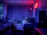A dimly lit stage setup in a bar or club featuring a drum kit, microphones, amplifiers, and a keyboard. The backdrop includes closed curtains and a disco ball. The walls are textured with a neon red light shaped like an animal and a sign reading 'Save Water, Drink Beer.' Seating and small tables with drinks are visible in the foreground.