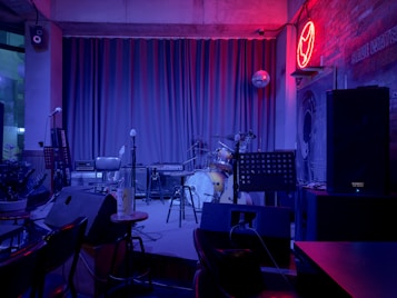 A dimly lit stage setup in a bar or club featuring a drum kit, microphones, amplifiers, and a keyboard. The backdrop includes closed curtains and a disco ball. The walls are textured with a neon red light shaped like an animal and a sign reading 'Save Water, Drink Beer.' Seating and small tables with drinks are visible in the foreground.