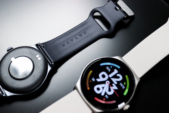 A smartwatch with a round face displaying digital time and colorful circular design elements on a black dial. The watch band appears to be made of silicone, with one side shown face up and the other displaying the underside with a clasp mechanism.