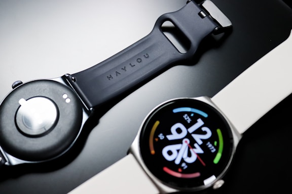 A smartwatch with a round face displaying digital time and colorful circular design elements on a black dial. The watch band appears to be made of silicone, with one side shown face up and the other displaying the underside with a clasp mechanism.