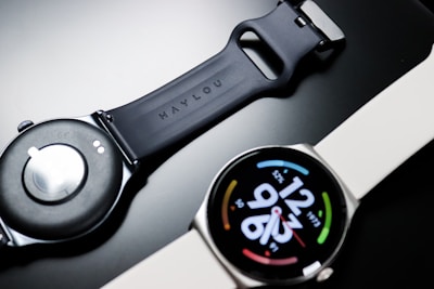 A smartwatch with a round face displaying digital time and colorful circular design elements on a black dial. The watch band appears to be made of silicone, with one side shown face up and the other displaying the underside with a clasp mechanism.