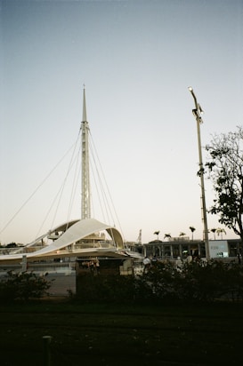 A modern architectural structure with a tall spire and tension cables, resembling a tent or sail. Surrounding the structure are people walking and some greenery with trees. The sky is clear with a light gradient, transitioning from a light blue at the top to softer tones near the horizon.