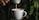 A stream of coffee flows from a small coffee machine into a white ceramic cup. The background features blurred green leaves, suggesting the presence of plants. The lighting is dim, creating a cozy and intimate ambiance.