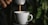 A stream of coffee flows from a small coffee machine into a white ceramic cup. The background features blurred green leaves, suggesting the presence of plants. The lighting is dim, creating a cozy and intimate ambiance.