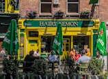 A lively street scene featuring a pub named 'Hairy Lemon'. The entrance is painted yellow and green and has hanging flower pots above. There are green umbrellas with 'Heineken' logos shading outdoor tables where people are sitting and talking. The windows on the second floor have flower boxes and there are signs for food and drinks.