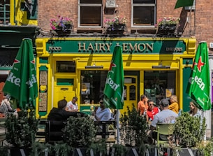 A lively street scene featuring a pub named 'Hairy Lemon'. The entrance is painted yellow and green and has hanging flower pots above. There are green umbrellas with 'Heineken' logos shading outdoor tables where people are sitting and talking. The windows on the second floor have flower boxes and there are signs for food and drinks.