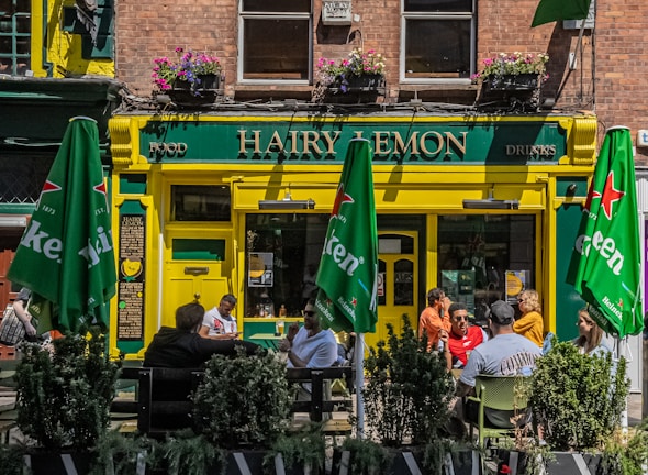 A lively street scene featuring a pub named 'Hairy Lemon'. The entrance is painted yellow and green and has hanging flower pots above. There are green umbrellas with 'Heineken' logos shading outdoor tables where people are sitting and talking. The windows on the second floor have flower boxes and there are signs for food and drinks.