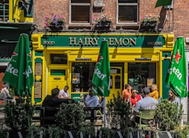 A lively street scene featuring a pub named 'Hairy Lemon'. The entrance is painted yellow and green and has hanging flower pots above. There are green umbrellas with 'Heineken' logos shading outdoor tables where people are sitting and talking. The windows on the second floor have flower boxes and there are signs for food and drinks.