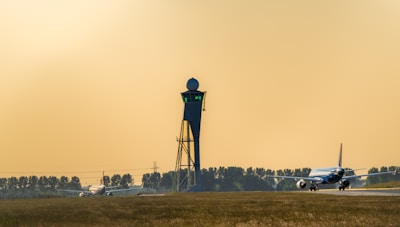 A control tower stands prominently on the airport runway with two airplanes visible, one on the taxiway and the other preparing for takeoff. The background features a row of trees and a clear, warm sky.