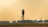 A control tower stands prominently on the airport runway with two airplanes visible, one on the taxiway and the other preparing for takeoff. The background features a row of trees and a clear, warm sky.