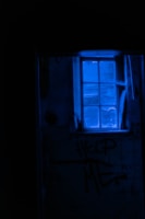 A dimly lit room with a blue-tinted window casting an eerie glow. The words 'HELP ME' are scrawled on the wall beneath the window, amidst a shadowy and neglected environment.