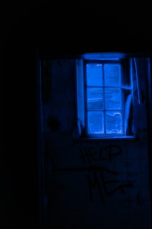 A dimly lit room with a blue-tinted window casting an eerie glow. The words 'HELP ME' are scrawled on the wall beneath the window, amidst a shadowy and neglected environment.
