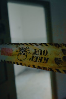 A piece of yellow caution tape with black lettering and a skull and crossbones symbol stretches across a dimly lit doorway. The words 'KEEP OUT' are prominently printed in bold letters.