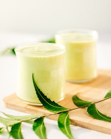 Two glasses filled with a creamy, light green smoothie are placed on a wooden board. Fresh green leaves, likely from a neem plant, are artfully arranged around the glasses, adding a natural and fresh element.