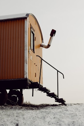 A small, elevated, yellow mobile structure with a rounded roof and metal chimney pipe is situated on a snowy ground. The structure is equipped with an external staircase, leading up to a door, and has visible wheels underneath.