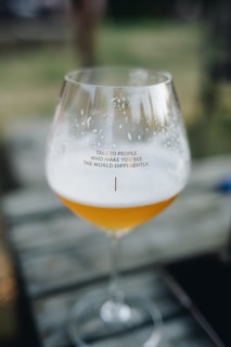 A clear glass goblet, partially filled with a frothy amber liquid, rests on a wooden surface. The text 'Talk to people who make you see the world differently' is printed on the glass. The background is blurred, suggesting an outdoor setting with muted earthy tones.