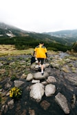 A hiker wearing a bright Traildry jacket crossing a rocky mountain stream under cloudy skies.
