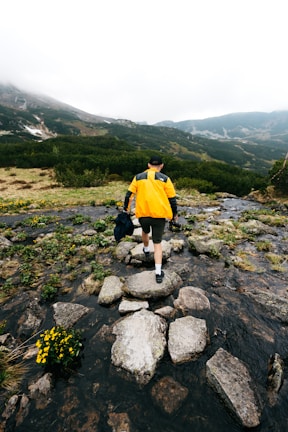 A hiker wearing a bright Traildry jacket crossing a rocky mountain stream under cloudy skies.