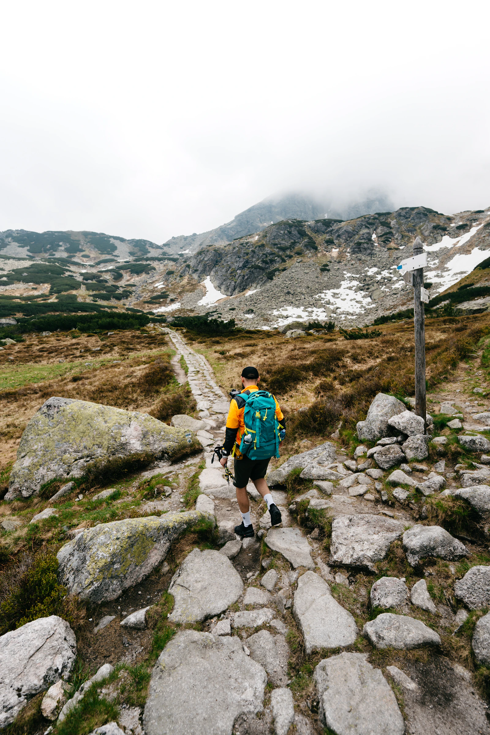 Photograph of A hiker climbing a rocky mountain path beside old snow while fog obscures the ridge.