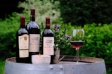 Three bottles of red wine are placed on a wooden barrel outdoors. A glass of red wine is partially filled and placed near the bottles. A small bowl with an orange substance and a potted plant with purple flowers are also present on the barrel. Lush green foliage in the background suggests a garden setting.